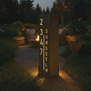 May include: A rectangular, dark metal address sign with the numbers 2347 and the word "SUNSET" cut out and illuminated. The sign is lit from within, casting a warm glow on the surrounding patio and lawn at dusk.