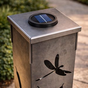 May include: A solar-powered outdoor light fixture with a square, metallic gray body. The design features a dragonfly and cattail cutouts. The top has a black solar panel. This decorative light is suitable for gardens and pathways.