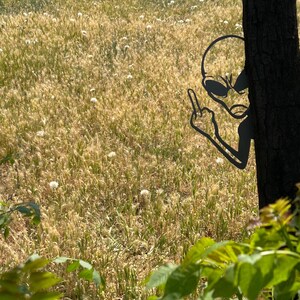 May include: A black metal alien silhouette giving the middle finger, positioned behind a tree trunk. The background features a field of tall, dry grass and dandelions, with green foliage in the foreground. The alien's design includes large oval eyes and a slender head.