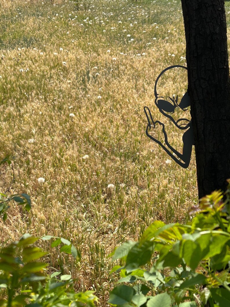 May include: A black metal alien silhouette giving the middle finger, positioned behind a tree trunk. The background features a field of tall, dry grass and dandelions, with green foliage in the foreground. The alien's design includes large oval eyes and a slender head.