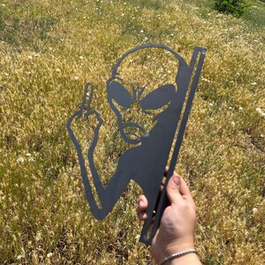 May include: A black metal alien silhouette with large eyes and a raised middle finger. The alien is held in a hand against a background of dry grass and wildflowers. The metal cutout has a vertical support.