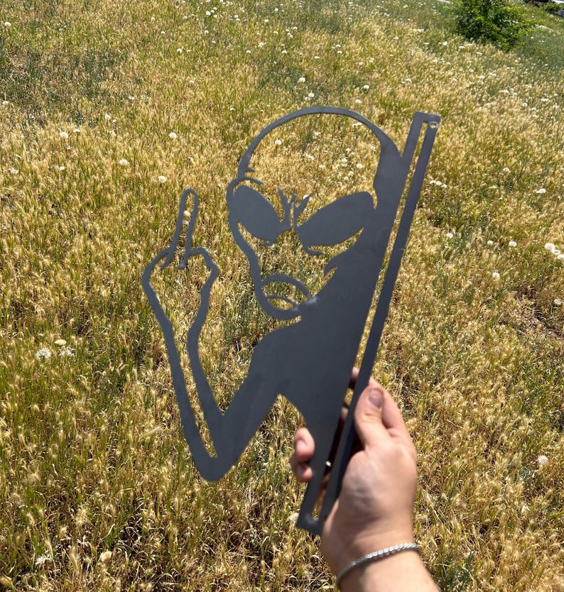 May include: A black metal alien silhouette with large eyes and a raised middle finger. The alien is held in a hand against a background of dry grass and wildflowers. The metal cutout has a vertical support.
