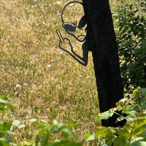 May include: A black metal alien silhouette giving the middle finger, attached to a tree trunk. The alien is in a field of tall grass and wildflowers, with green foliage in the foreground.