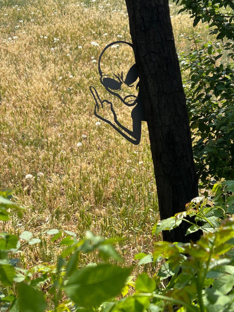 May include: A black metal alien silhouette giving the middle finger, attached to a tree trunk. The alien is in a field of tall grass and wildflowers, with green foliage in the foreground.