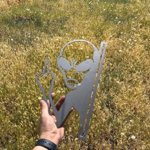 May include: A metal alien silhouette with a raised middle finger. The alien has large eyes and a small mouth. The metal is a silver color. The alien is held in a hand against a background of dry grass.