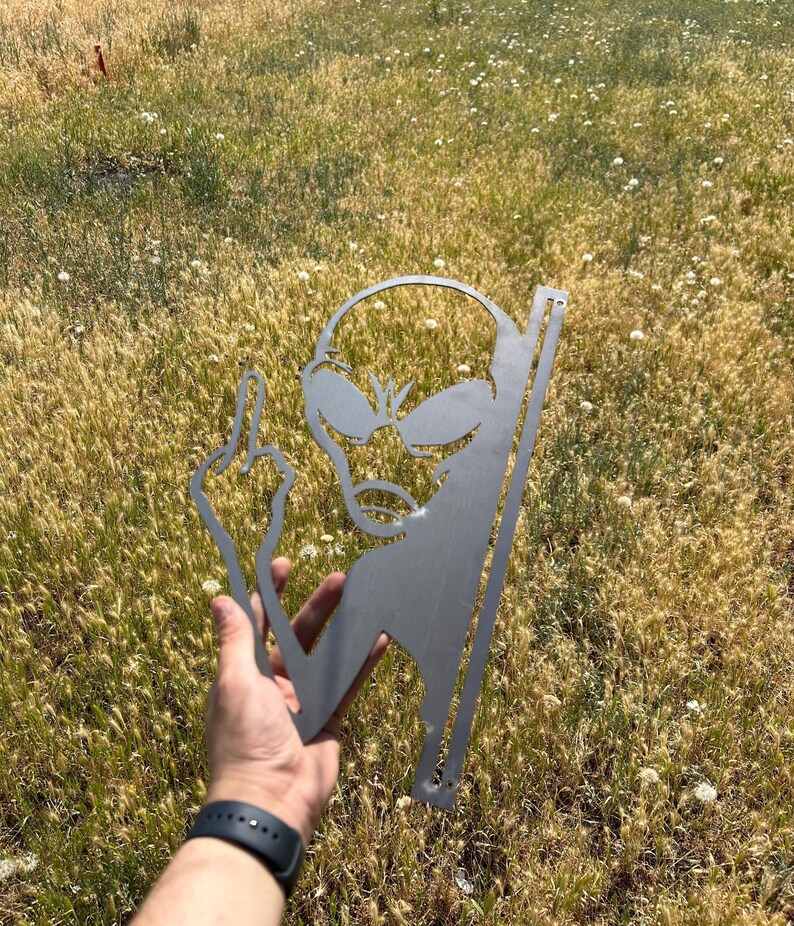 May include: A metal alien silhouette with a raised middle finger. The alien has large eyes and a small mouth. The metal is a silver color. The alien is held in a hand against a background of dry grass.