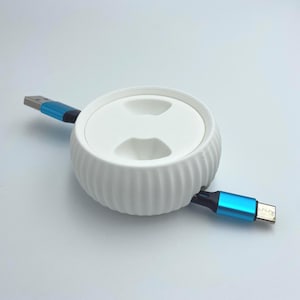 May include: A white cable organizer with a ribbed exterior and a flat top. Two cables with blue accents and USB connectors extend from the organizer. The product is designed to manage and store cables neatly.