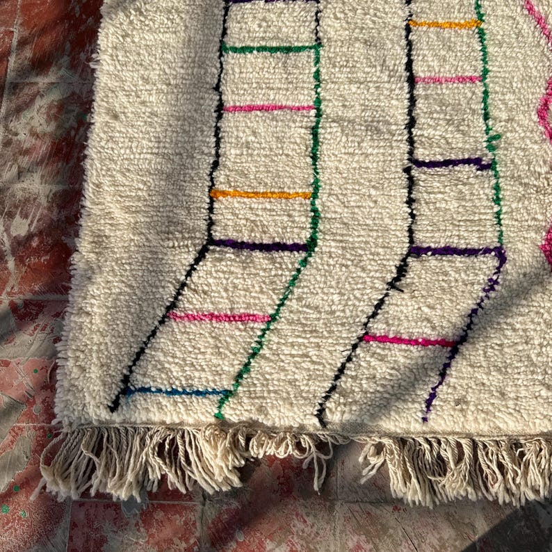Puede incluir: Alfombra texturizada de color crema con un dise&ntilde;o geom&eacute;trico. La alfombra presenta l&iacute;neas negras, verdes, rosas, amarillas y moradas, con flecos en el borde inferior. La alfombra est&aacute; colocada sobre una superficie de ladrillo rojo.