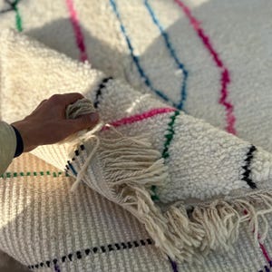 Puede incluir: Una alfombra crema texturizada con rayas coloridas en rosa, azul y verde. La alfombra tiene un detalle de flecos en un extremo. La alfombra est&aacute; doblada, revelando la parte inferior y una mano que sostiene el borde.