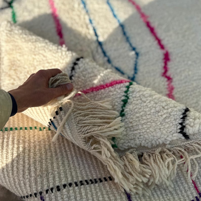 Puede incluir: Una alfombra crema texturizada con rayas coloridas en rosa, azul y verde. La alfombra tiene un detalle de flecos en un extremo. La alfombra est&aacute; doblada, revelando la parte inferior y una mano que sostiene el borde.