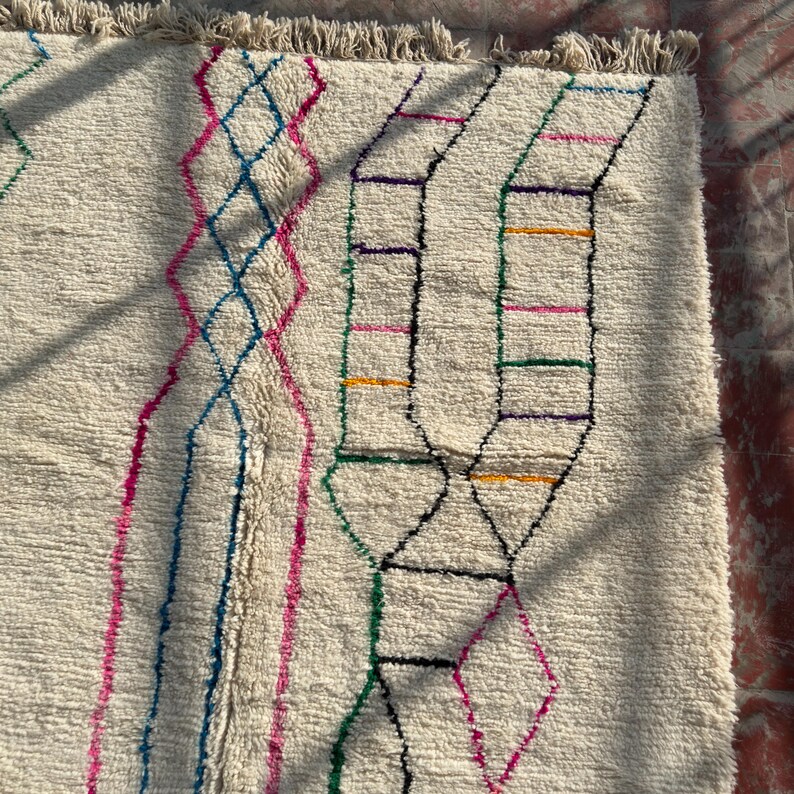 Puede incluir: Alfombra texturizada de color crema con patrones geom&eacute;tricos coloridos. El dise&ntilde;o incluye formas de diamante y rectangulares en rosa, azul, verde, amarillo y morado. La alfombra tiene un borde con flecos.