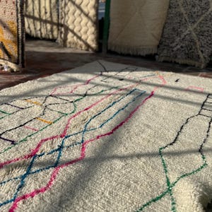 Puede incluir: Una alfombra de color crema con un dise&ntilde;o geom&eacute;trico en l&iacute;neas rosas, azules, verdes y negras. La alfombra tiene una superficie texturizada y se muestra con luz natural, con otras alfombras visibles en el fondo.