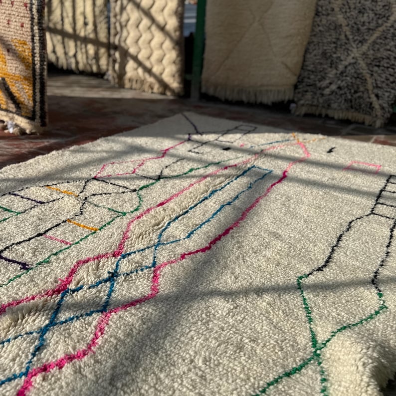 Puede incluir: Una alfombra de color crema con un dise&ntilde;o geom&eacute;trico en l&iacute;neas rosas, azules, verdes y negras. La alfombra tiene una superficie texturizada y se muestra con luz natural, con otras alfombras visibles en el fondo.
