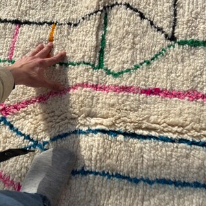 Puede incluir: Alfombra texturizada de color crema con un dise&ntilde;o geom&eacute;trico colorido. La alfombra presenta l&iacute;neas en rosa, verde, azul, amarillo y negro. La textura de la alfombra es visible, con una mano y un pie parcialmente visibles.