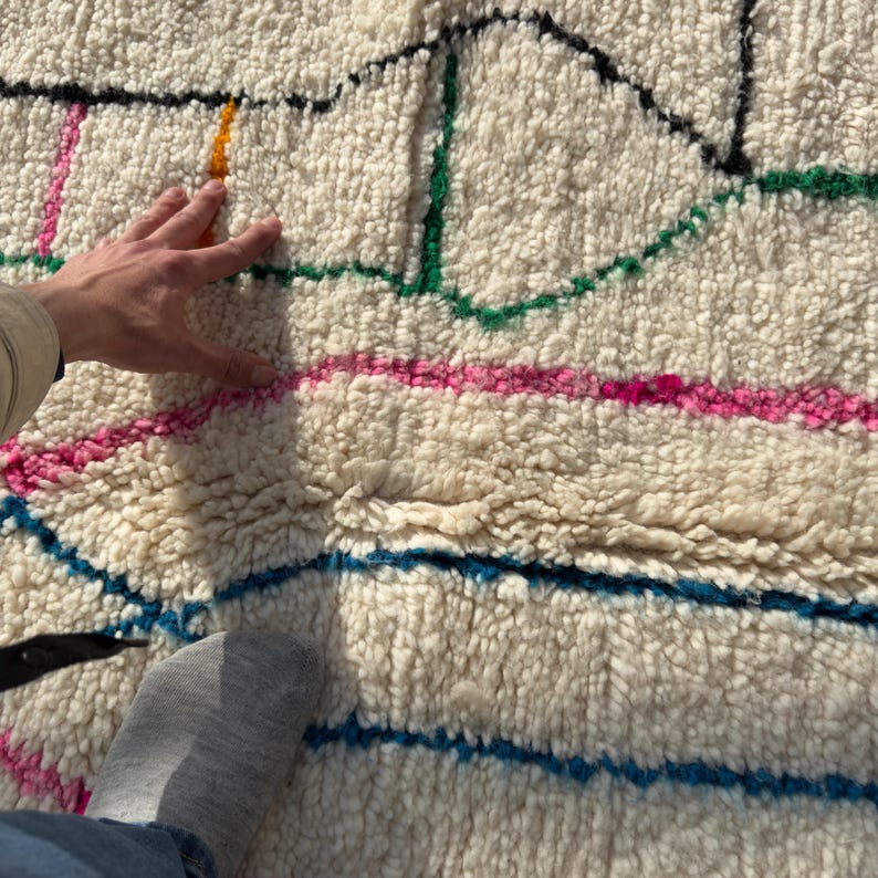 Puede incluir: Alfombra texturizada de color crema con un dise&ntilde;o geom&eacute;trico colorido. La alfombra presenta l&iacute;neas en rosa, verde, azul, amarillo y negro. La textura de la alfombra es visible, con una mano y un pie parcialmente visibles.