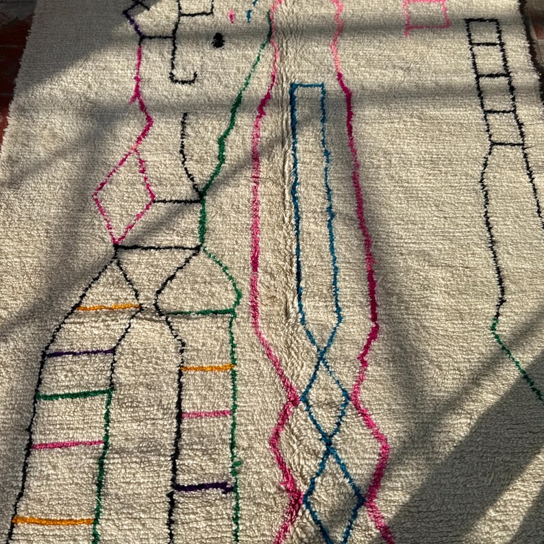 Puede incluir: Alfombra de color crema con un dise&ntilde;o geom&eacute;trico. La alfombra presenta un patr&oacute;n de l&iacute;neas y formas negras, rosas, verdes, azules y amarillas. El dise&ntilde;o incluye formas de diamante, l&iacute;neas verticales y otras formas abstractas. La alfombra tiene una superficie texturizada.