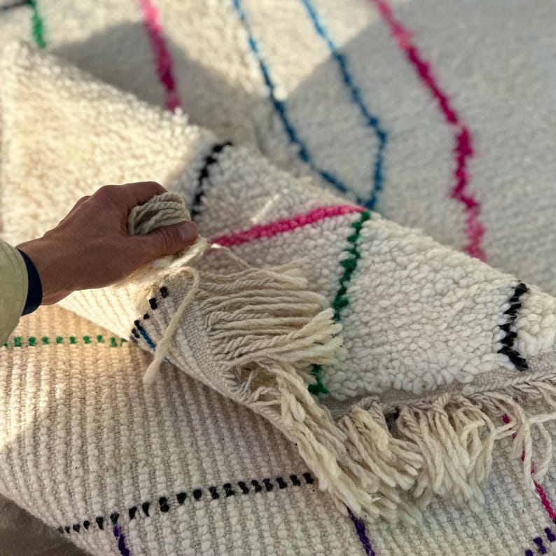 Puede incluir: Alfombra texturizada de color crema con rayas coloridas en rosa, verde, azul y morado. La alfombra tiene un detalle de flecos en el borde y un patr&oacute;n de cuadr&iacute;cula. La alfombra est&aacute; doblada, revelando la parte inferior.