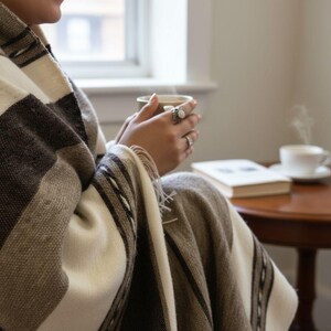 May include: A striped blanket in shades of brown, beige, and white. Someone is holding a warm drink in a ceramic mug. A book and a cup of coffee are on a small table, suggesting a comfortable, relaxed setting.
