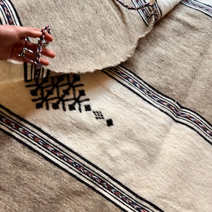 Puede incluir: Textil tejido beige y blanco con patrones geométricos negros y bordes a rayas. El textil tiene un borde con flecos y está parcialmente enrollado, revelando la parte inferior. El diseño incluye detalles en negro, rojo y azul.