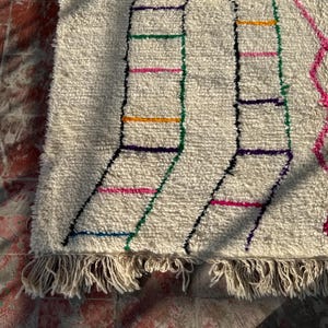 Puede incluir: Alfombra texturizada de color crema con un dise&ntilde;o geom&eacute;trico. La alfombra presenta l&iacute;neas negras que forman formas de escalera, acentuadas con rayas horizontales coloridas en rosa, verde, naranja y morado. La alfombra tiene un borde con flecos.