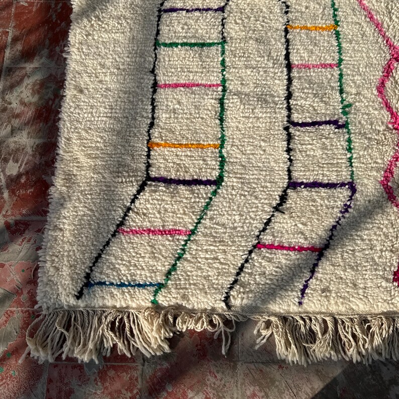 Puede incluir: Alfombra texturizada de color crema con un dise&ntilde;o geom&eacute;trico. La alfombra presenta l&iacute;neas negras que forman formas de escalera, acentuadas con rayas horizontales coloridas en rosa, verde, naranja y morado. La alfombra tiene un borde con flecos.
