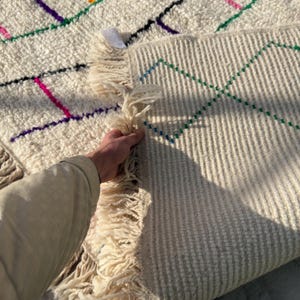 Puede incluir: Alfombra color crema con dise&ntilde;os geom&eacute;tricos coloridos. La alfombra presenta una superficie texturizada y flecos en los bordes. El dise&ntilde;o incluye l&iacute;neas en rosa, morado, verde y negro.