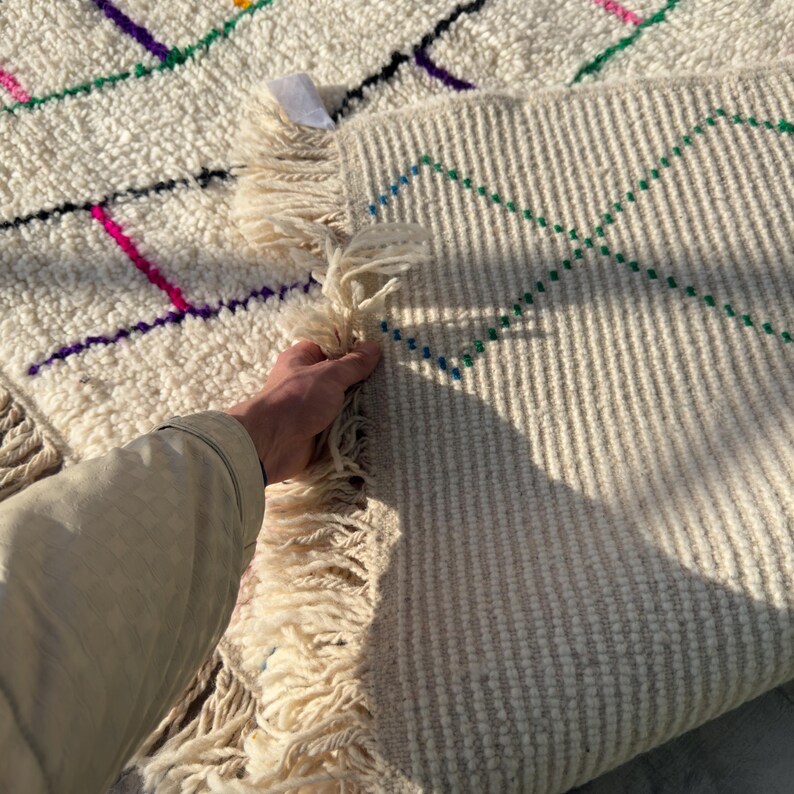 Puede incluir: Alfombra color crema con dise&ntilde;os geom&eacute;tricos coloridos. La alfombra presenta una superficie texturizada y flecos en los bordes. El dise&ntilde;o incluye l&iacute;neas en rosa, morado, verde y negro.