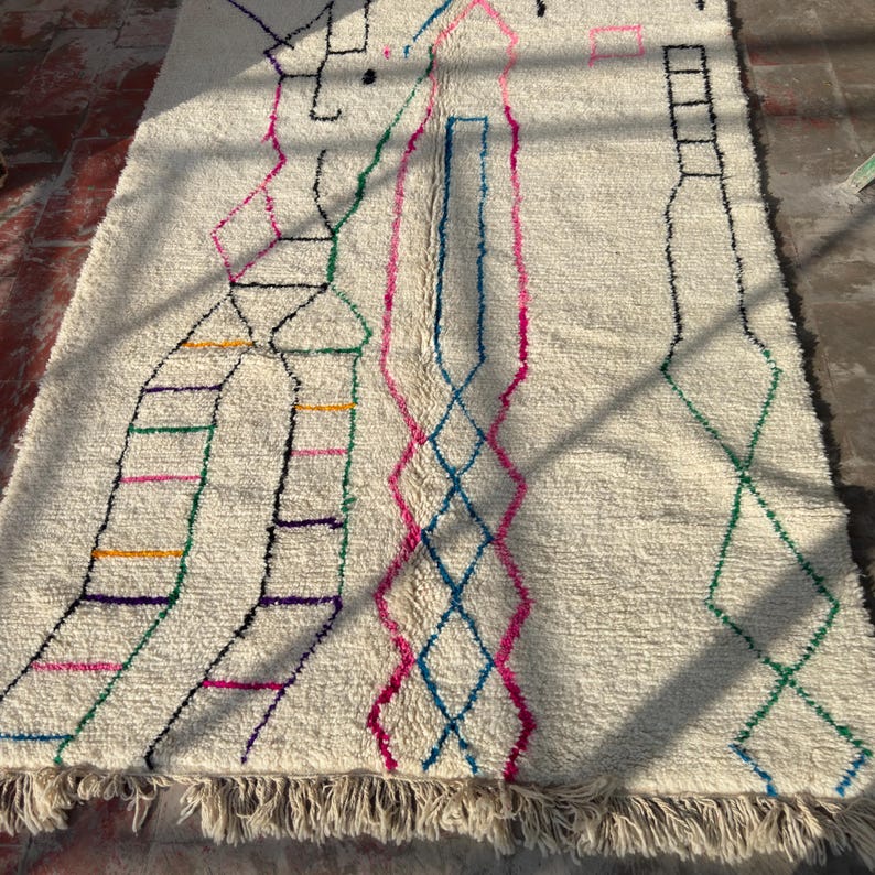 Puede incluir: Alfombra texturizada de color crema con un dise&ntilde;o geom&eacute;trico. La alfombra presenta acentos coloridos en rosa, azul, verde y amarillo. El dise&ntilde;o incluye formas de diamante y rectangulares, con un borde con flecos.