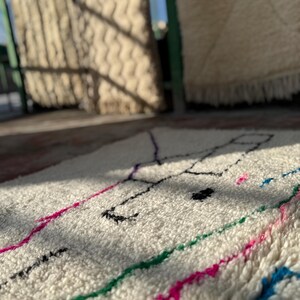 Puede incluir: Alfombra de color crema con un dise&ntilde;o geom&eacute;trico. La alfombra presenta un patr&oacute;n de l&iacute;neas negras, moradas, rosas, verdes y azules. La alfombra est&aacute; hecha de un material suave y texturizado.