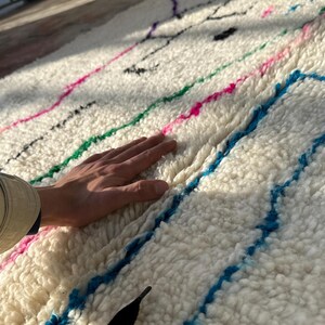 Puede incluir: Una alfombra blanca y texturizada con rayas coloridas en rosa, verde y azul. La alfombra tiene un pelo grueso y afelpado y una mano descansa sobre la superficie. La alfombra parece estar hecha de lana o un material similar.