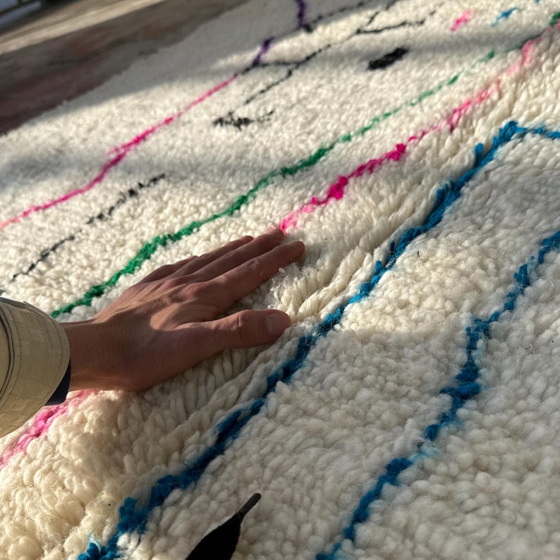 Puede incluir: Una alfombra blanca y texturizada con rayas coloridas en rosa, verde y azul. La alfombra tiene un pelo grueso y afelpado y una mano descansa sobre la superficie. La alfombra parece estar hecha de lana o un material similar.