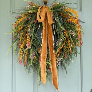 May include: A teardrop-shaped wreath featuring green, orange, and red foliage with small clusters of light-colored berries. A long, wide, mustard-yellow velvet ribbon with frayed edges is tied at the top, hanging down the center of the wreath.