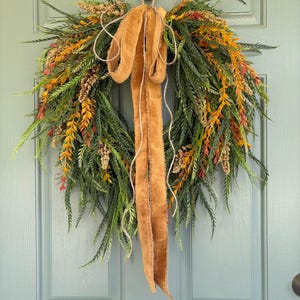May include: A fall-themed wreath with green, orange, and red foliage. A wide, tan velvet ribbon is tied in a bow at the top and hangs down. The wreath is hanging on a light blue door.