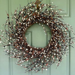 May include: A circular wreath constructed from thin, dark brown intertwined twigs. The wreath is densely decorated with numerous small, round berries in contrasting colors of matte white and glossy black, interspersed throughout the branch structure.