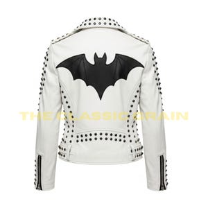 Puede incluir: Chaqueta de cuero blanca con un emblema de murciélago negro en la espalda, tachonada con remaches plateados. Diseño clásico de chaqueta de motorista con cuello de solapa, detalles de cremallera y el texto "THE CLASSIC RAIN".