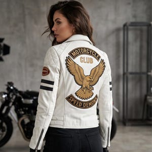 Könnte beinhalten: Eine weiße Motorradjacke aus Leder mit einem großen gestickten Aufnäher auf dem Rücken, der einen goldenen Adler und den Text MOTORCYCLE CLUB SPEED DEMONS zeigt. Die Jacke hat gesteppte Schulterpartien, schwarze Ärmelstreifen und Metallreißverschlüsse.
