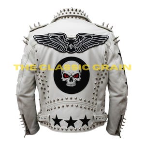 Könnte beinhalten: Weiße Lederjacke mit silbernen Stacheln auf Schultern und Kragen. Verfügt über ein schwarz-weißes geflügeltes Emblem und eine Totenkopf-Grafik auf dem Rücken. Enthält drei schwarze Sterne und den Text "THE CLASSIC GRAIN".