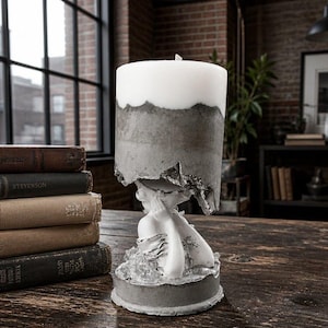 May include: A cylindrical white candle with a textured gray concrete base and midsection, featuring a sculpted white figure with hands near the face at the bottom. The candle rests on a circular gray concrete pedestal with silver accents.