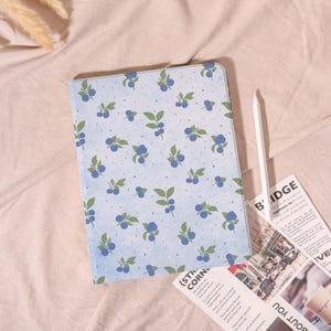 May include: A light blue tablet case with a repeating pattern of blueberries and green leaves. A white stylus and a newspaper are next to the case. The case is on a pink surface.