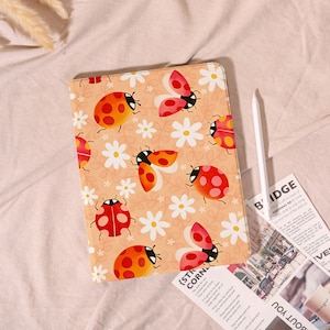 May include: A tablet case with a repeating pattern of red and orange ladybugs and white daisies on a peach background. A white stylus and a newspaper are next to the case.