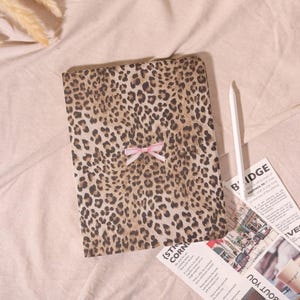 May include: A notebook with a leopard print cover and a small pink bow. The notebook is on a pink surface with a white stylus and a newspaper. The leopard print has brown and black spots on a tan background.