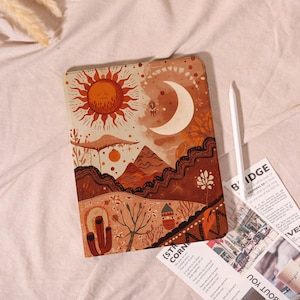 May include: Tablet case with a desert landscape design featuring a sun, moon, mountains, and cacti in shades of orange, brown, and cream. A white stylus and printed papers are next to the case.