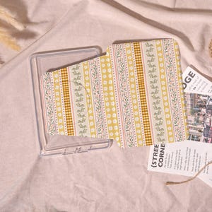 May include: A clear phone case with a patterned insert. The insert features a design with yellow, white, and pink floral and striped patterns. The text "{STREE CORNE" is visible on a small card.