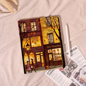 May include: A rectangular notebook featuring a vibrant illustration of two storefronts at dusk with warm yellow lighting, dark trees, and falling autumn leaves. The shop sign reads "Mare Boutlhks". A white stylus rests beside the notebook.