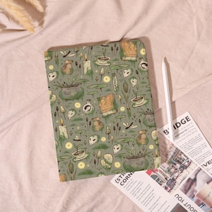 May include: A green tablet case featuring a repeating pattern of illustrations, including candles, potion bottles, and mushrooms. A white stylus is next to the case. The case is on a pink surface.