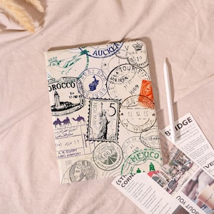 May include: A cream-coloured journal featuring a collage of travel-themed stamps, including "Morocco," "New York," and "Mexico." A white stylus and printed papers are next to the journal. The journal has a vintage aesthetic.