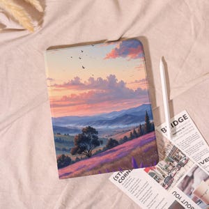 May include: A tablet case featuring a landscape painting of a sunset over rolling hills and fields of purple flowers. A white stylus and a newspaper are next to the case. The case has a smooth finish.