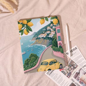 May include: A colorful tablet case featuring a coastal Italian scene with a yellow car driving along a road. The case has a vibrant illustration of lemons, buildings, and the sea. A white stylus and printed papers are next to the case.