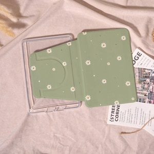 Könnte beinhalten: Eine hellgrüne Tablethülle mit einem Gänseblümchenmuster. Die Hülle ist geöffnet und zeigt das Innere und einen transparenten Kunststoffrahmen. Der Text "{STREET CORNER}" ist auf einem Papierstück zu sehen.