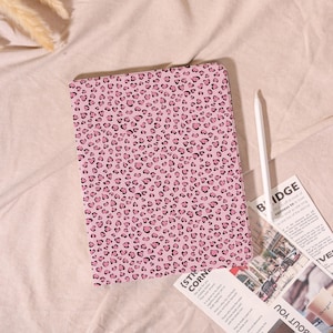 May include: Pink tablet case with a leopard print pattern of black hearts. A white stylus and a newspaper are next to the case. The case is on a pink fabric surface.